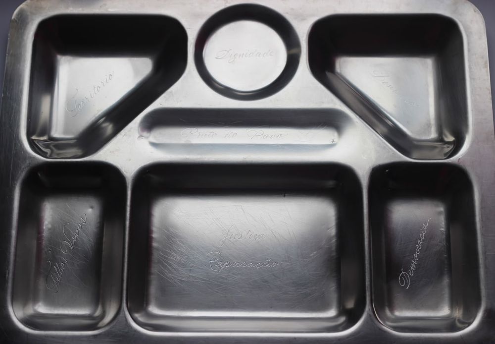Bandeja de metal de refeitório, com sete compartimentos e palavras gravadas em letra cursiva, vista de cima.