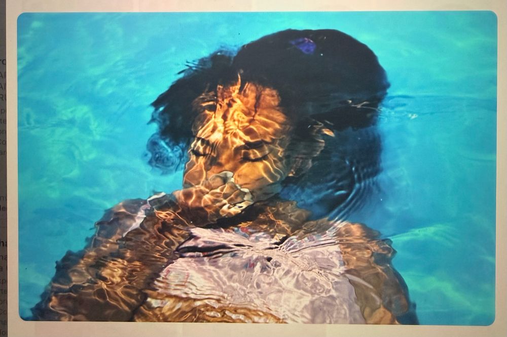 Fotografia de uma mulher quase submersa em água turquesa, de olhos fechados, com manchas douradas de luz sobre a pele.