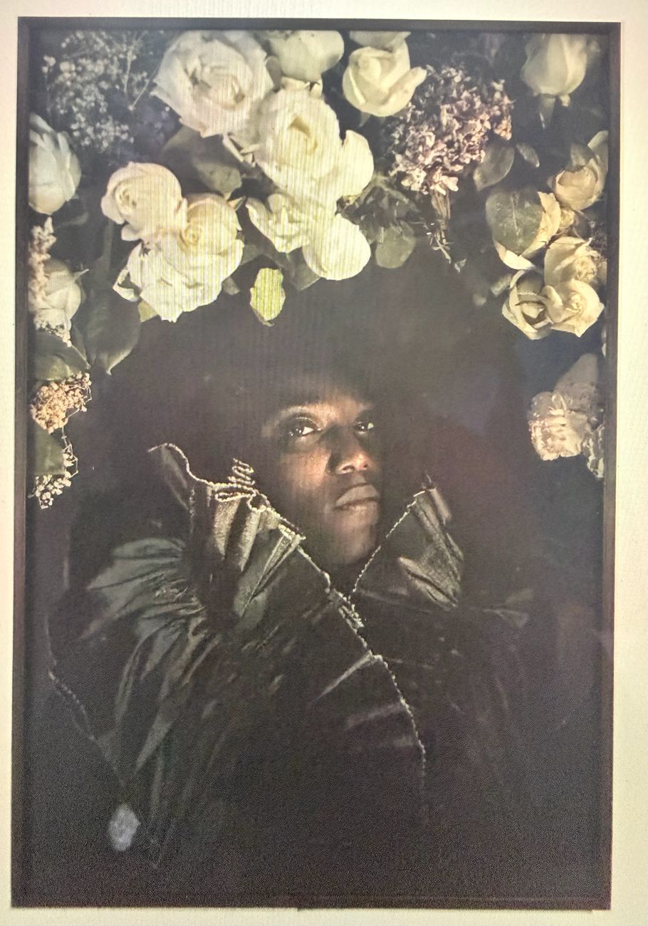 Retrato de uma mulher negra com grande coroa de rosas brancas e flores lilás, em tons escuros.